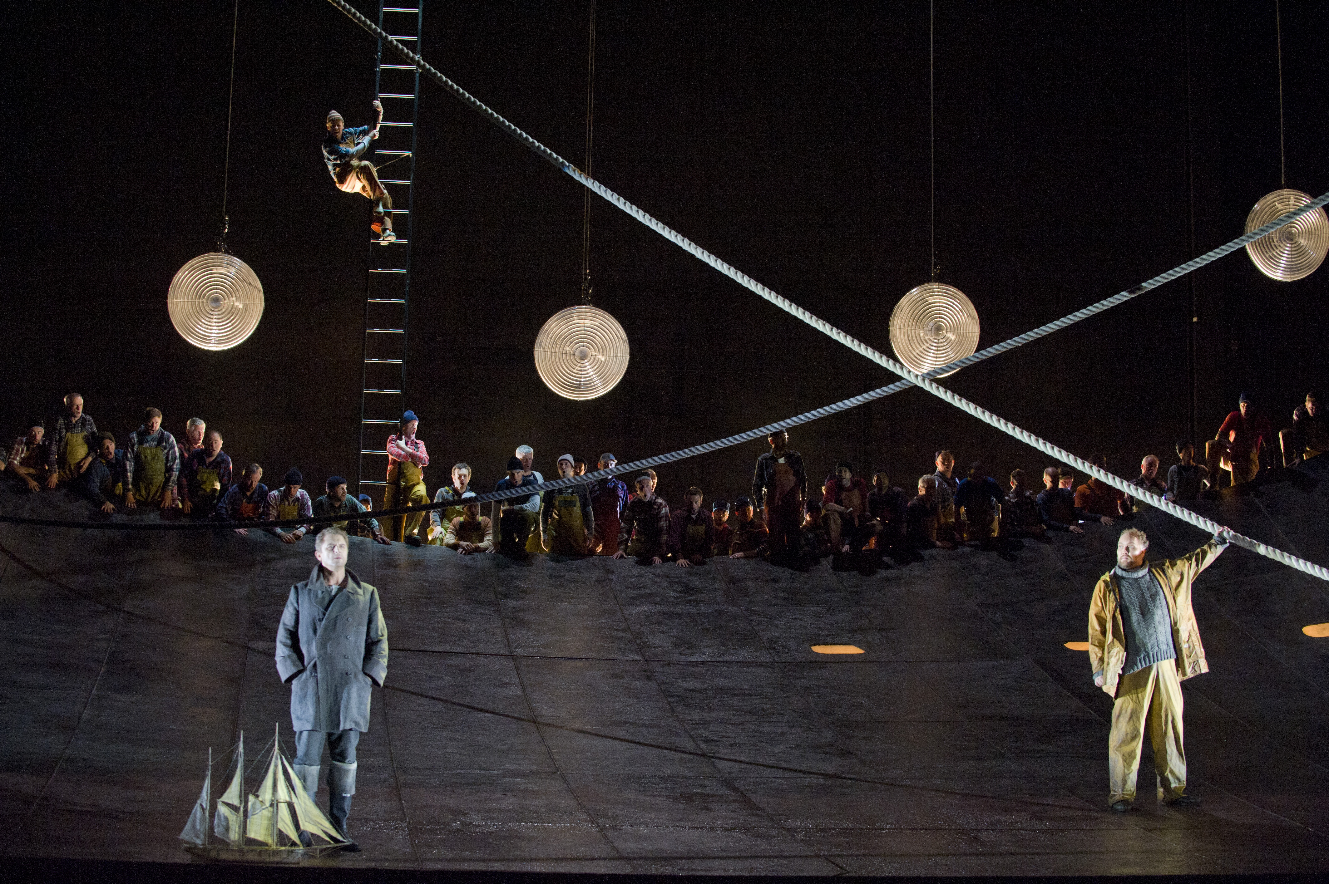 A man in grey overcoat stands next to a model ship. Another man in a beige stand the other side of the stage holding a sailors rope that is above his head. 