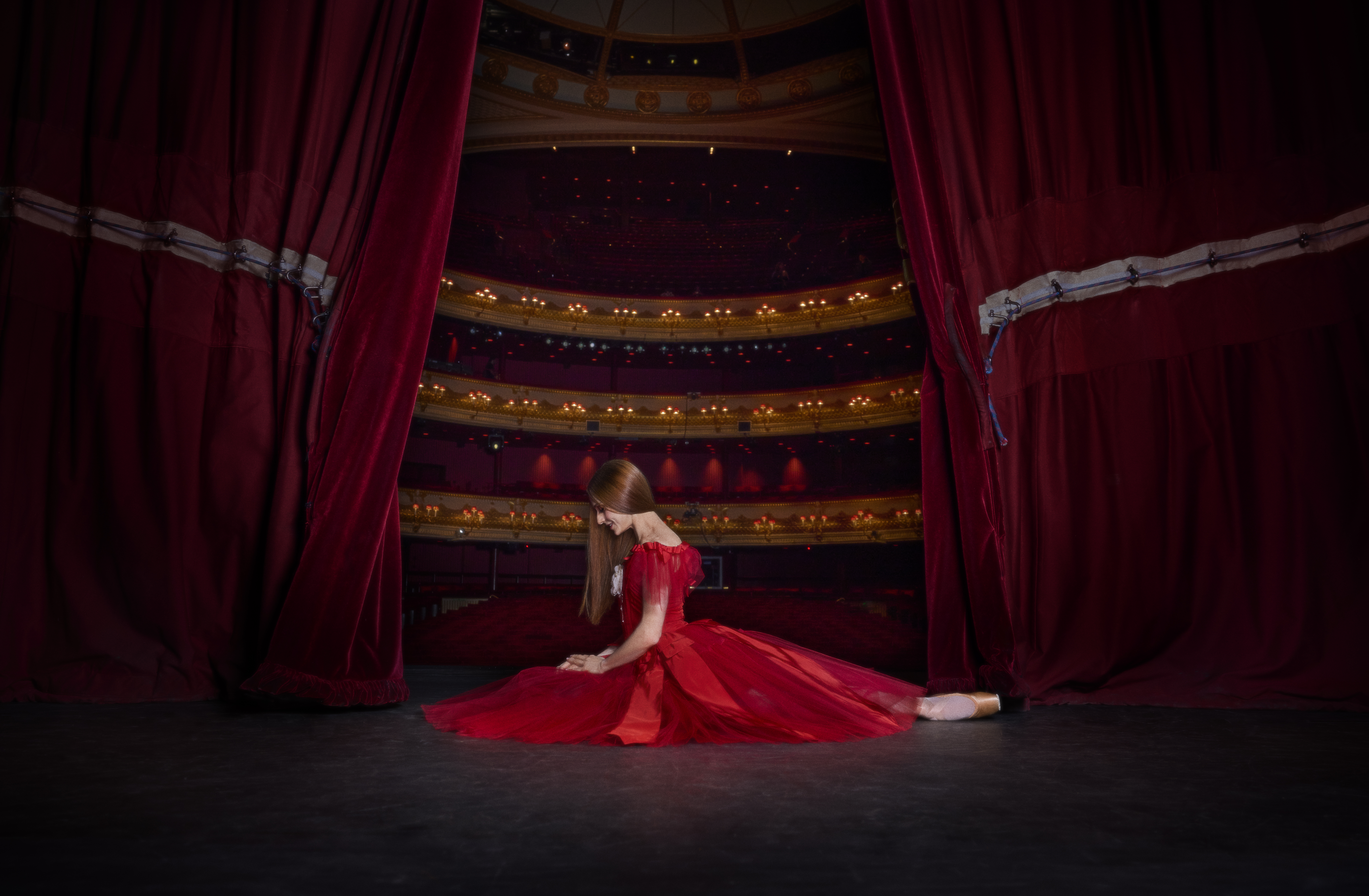 A ballerina kneels on the floor of a stage. She is wearing a red dress and her hair is long, dark and hanging by her face. Behind her the stage curtains are open slightly, revealing the lights and seats of the auditorium.