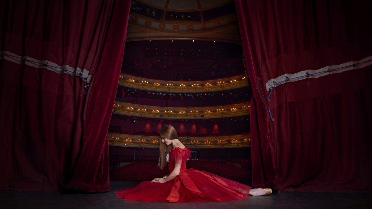 A ballerina kneels on the floor of a stage. She is wearing a red dress and her hair is long, dark and hanging by her face. Behind her the stage curtains are open slightly, revealing the lights and seats of the auditorium.