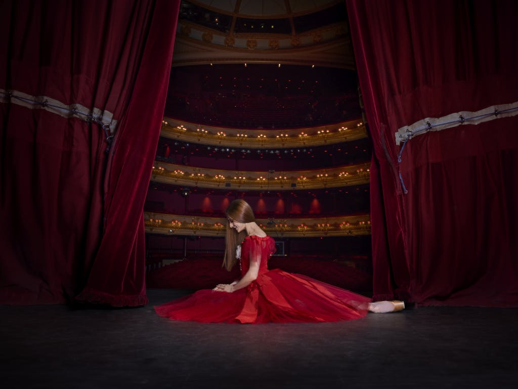 A ballerina kneels on the floor of a stage. She is wearing a red dress and her hair is long, dark and hanging by her face. Behind her the stage curtains are open slightly, revealing the lights and seats of the auditorium.