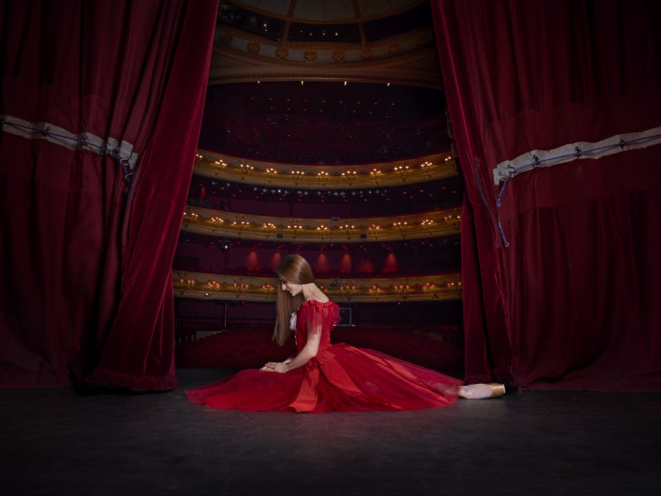 A ballerina kneels on the floor of a stage. She is wearing a red dress and her hair is long, dark and hanging by her face. Behind her the stage curtains are open slightly, revealing the lights and seats of the auditorium.