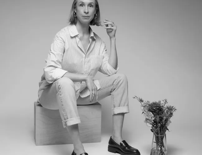 A woman with short hair sits on a box. There is a vase of flowers at her feet (2 of 5)