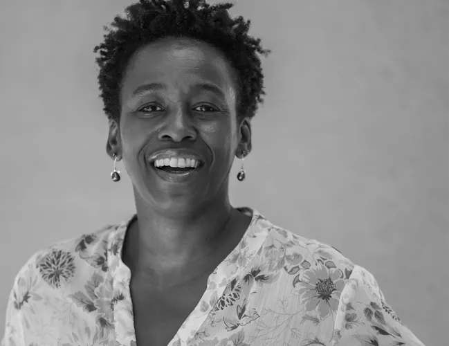 A black woman with medium-brown skin, brown eyes, and a slight, close-mouthed smile peers out at the viewer. Her hair is a short afro with twisted, uneven ends. She wears a v-neck top with a large floral pattern. (6 of 14)