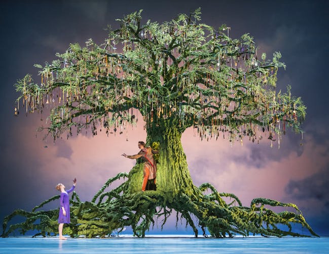 The Royal Ballet performing Winter's Tale at the Royal Opera House. A tall tree covered in green moss is decorated with hanging medallions. The sprawling roots reach across the stage. A blonde dancer in a purple dress holds her hand to a dancer in brown. The dancer is disappearing into a door in the trunk of the tree.