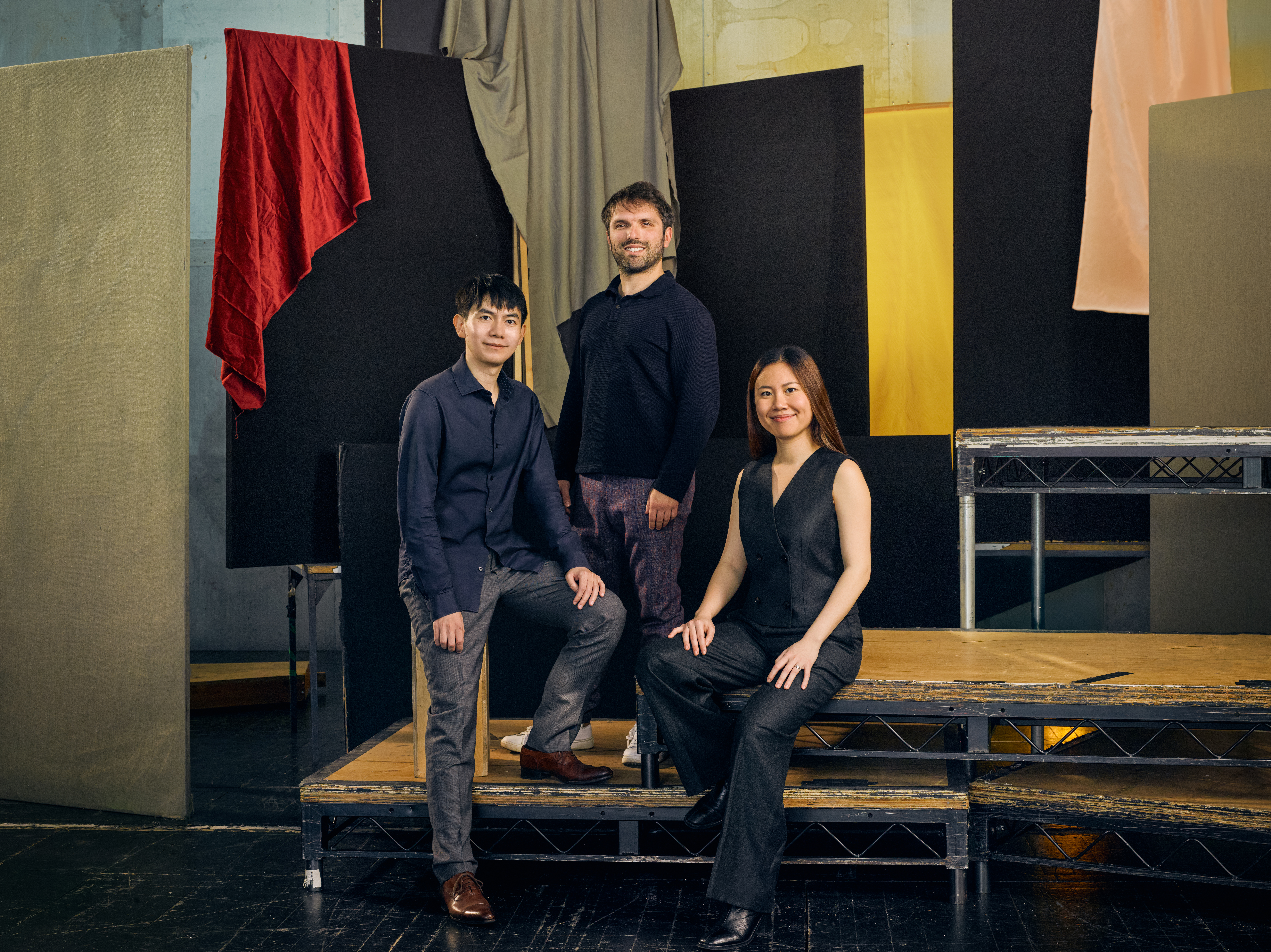 Jette Parker Artists Peggy Wu, Richard Fu and Giovanni Panizza stand in the centre of the photo. They are all wearing black. They stand on tiered stage pallets and there is a multi-coloured stage set behind them. 