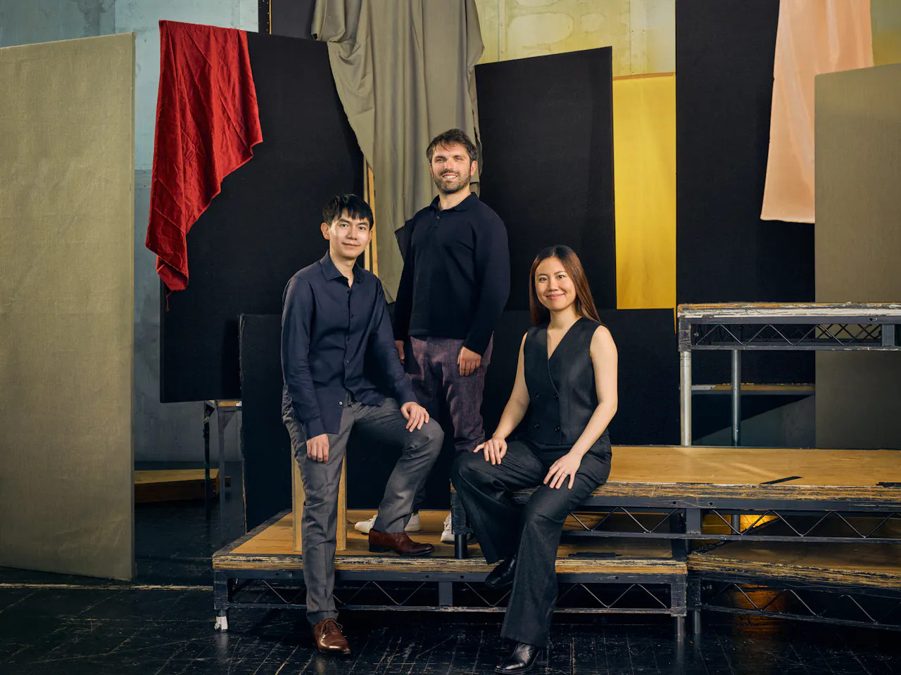Jette Parker Artists Peggy Wu, Richard Fu and Giovanni Panizza stand in the centre of the photo. They are all wearing black. They stand on tiered stage pallets and there is a multi-coloured stage set behind them.