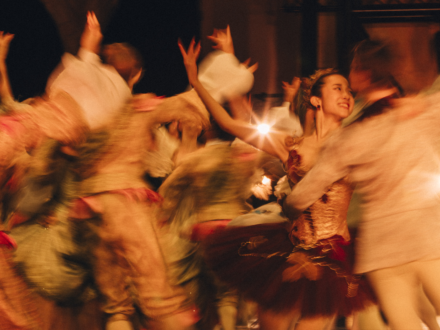 Several dancers wearing elaborate costumes dance close together with their arms raised. The image is artistically blurred to look like in motion. 