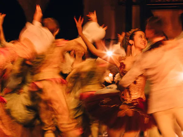 Several dancers wearing elaborate costumes dance close together with their arms raised. The image is artistically blurred to look like in motion.