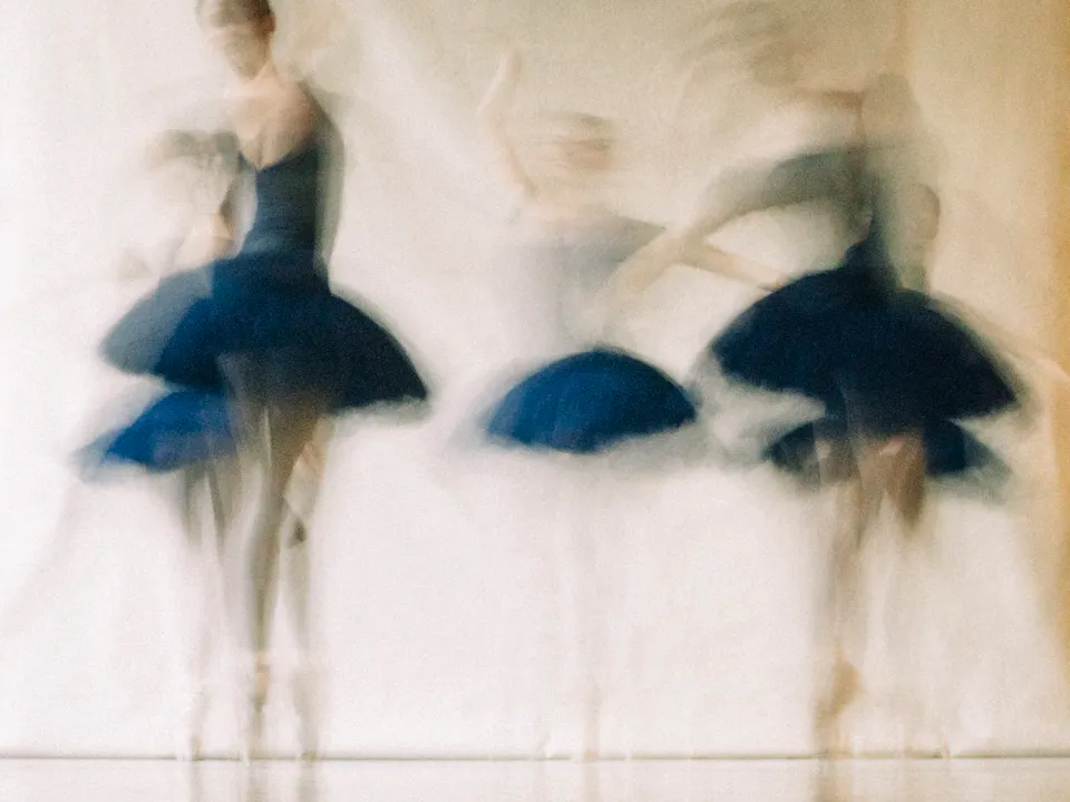 five ballet dancers wearing navy blue tutus stand on pointe with their arms upwards. The image is artistically blurred to look like in motion.
