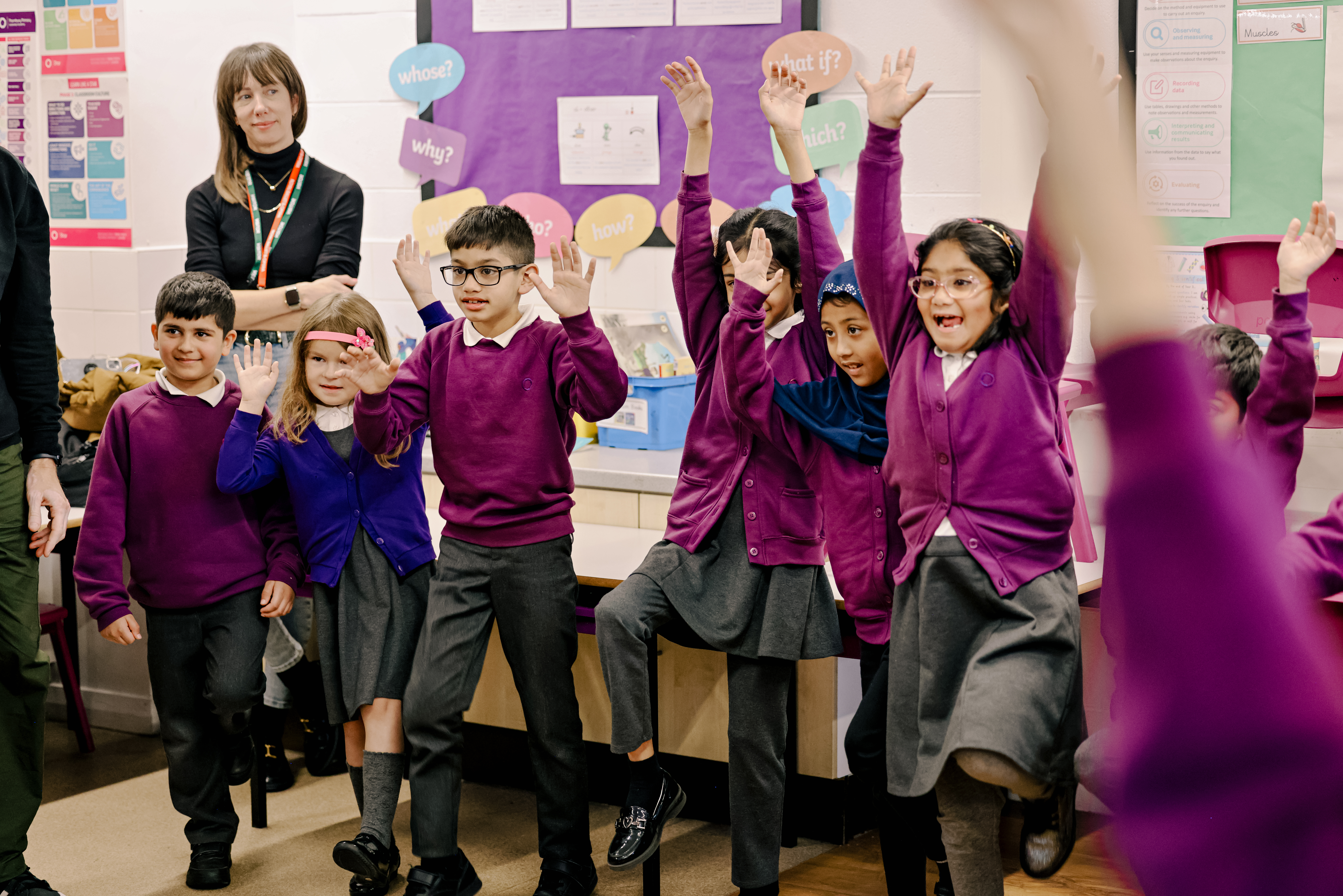 A group of children in purple jumpers and black trousers and skirts stand in a classroom with their arms raised. Behind them a woman with dark hair and a fringe is watching.
