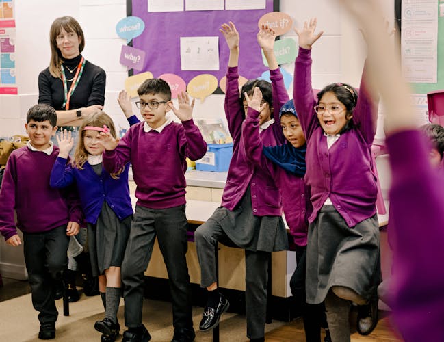 A group of children in purple jumpers and black trousers and skirts stand in a classroom with their arms raised. Behind them a woman with dark hair and a fringe is watching.