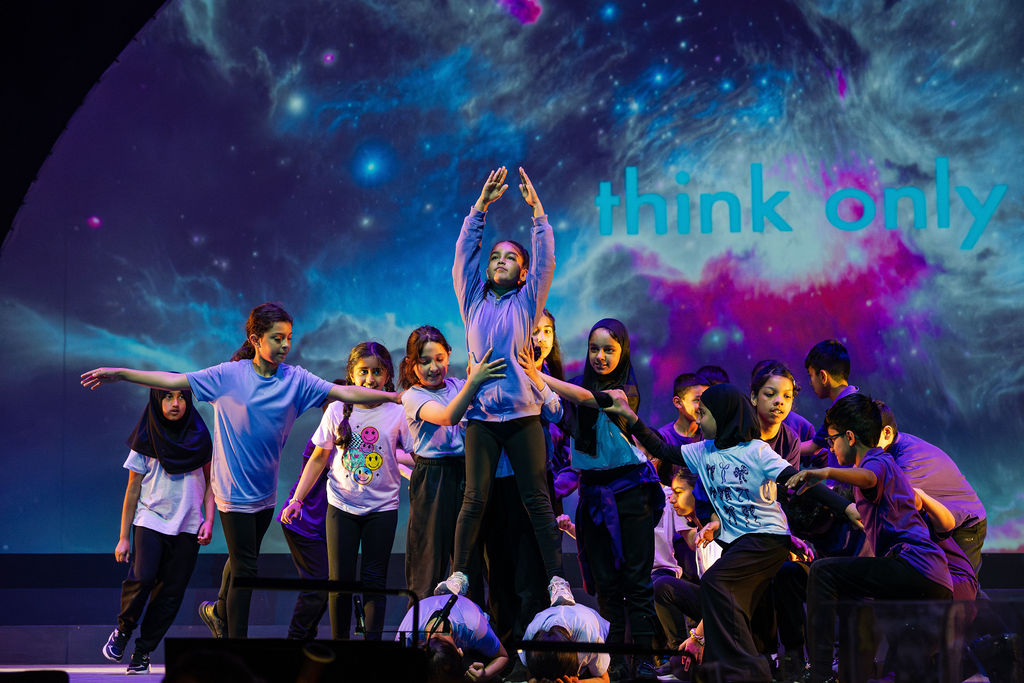 A group of children are performing on a stage. A girl balances with her hands in the air while other children support her, holding her waist on all sides. Behind them is a screen showing a space-like image.