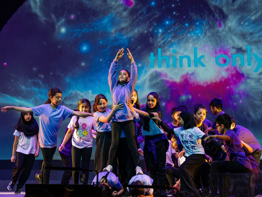 A group of children are performing on a stage. A girl balances with her hands in the air while other children support her, holding her waist on all sides. Behind them is a screen showing a space-like image.