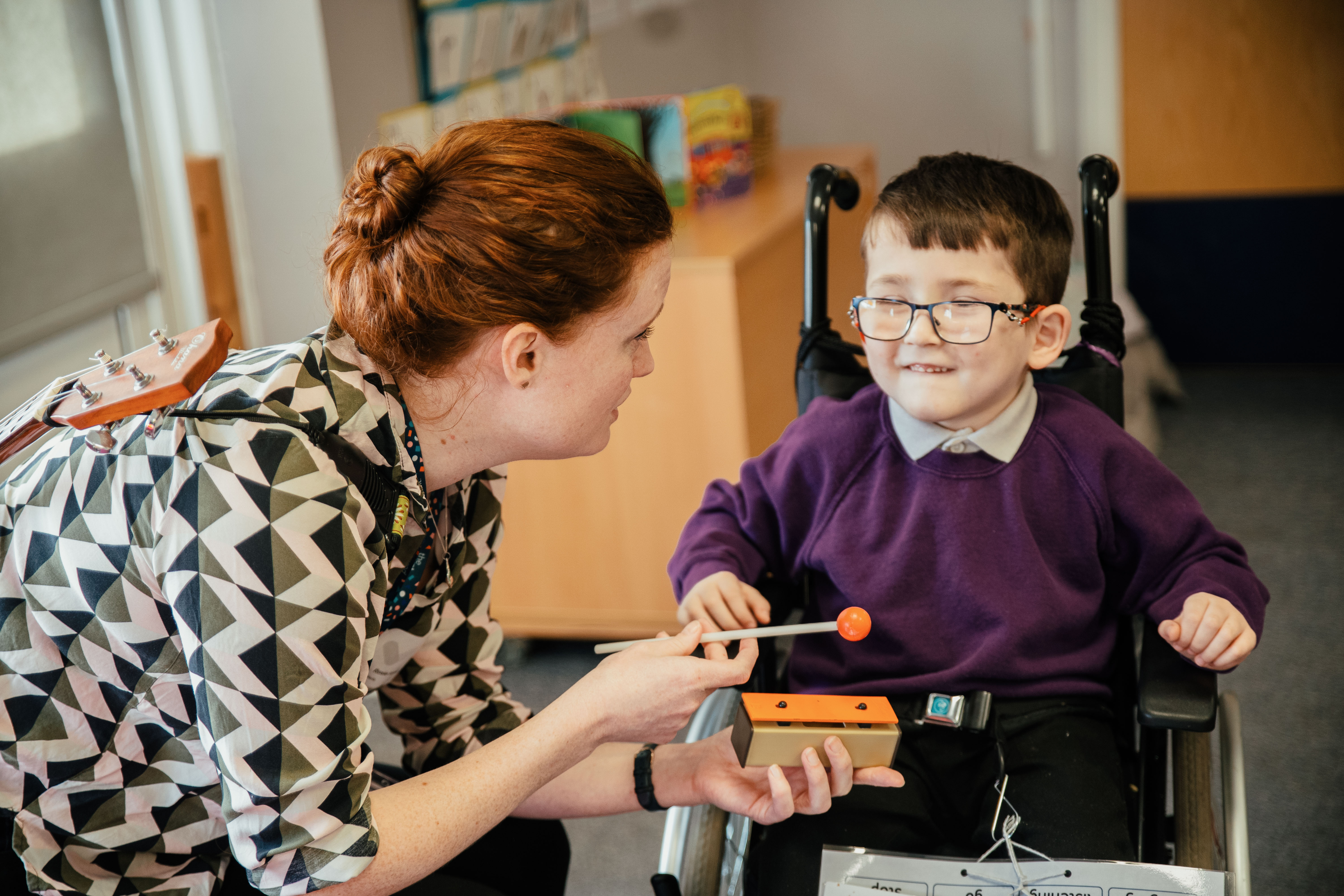 A woman with ginger hair and a shirt with geometric patterns kneels near a boy in a wheelchair. He is wearing a white polo neck, purple jumper and glasses. The woman is showing the boy a chime bar with a beater.