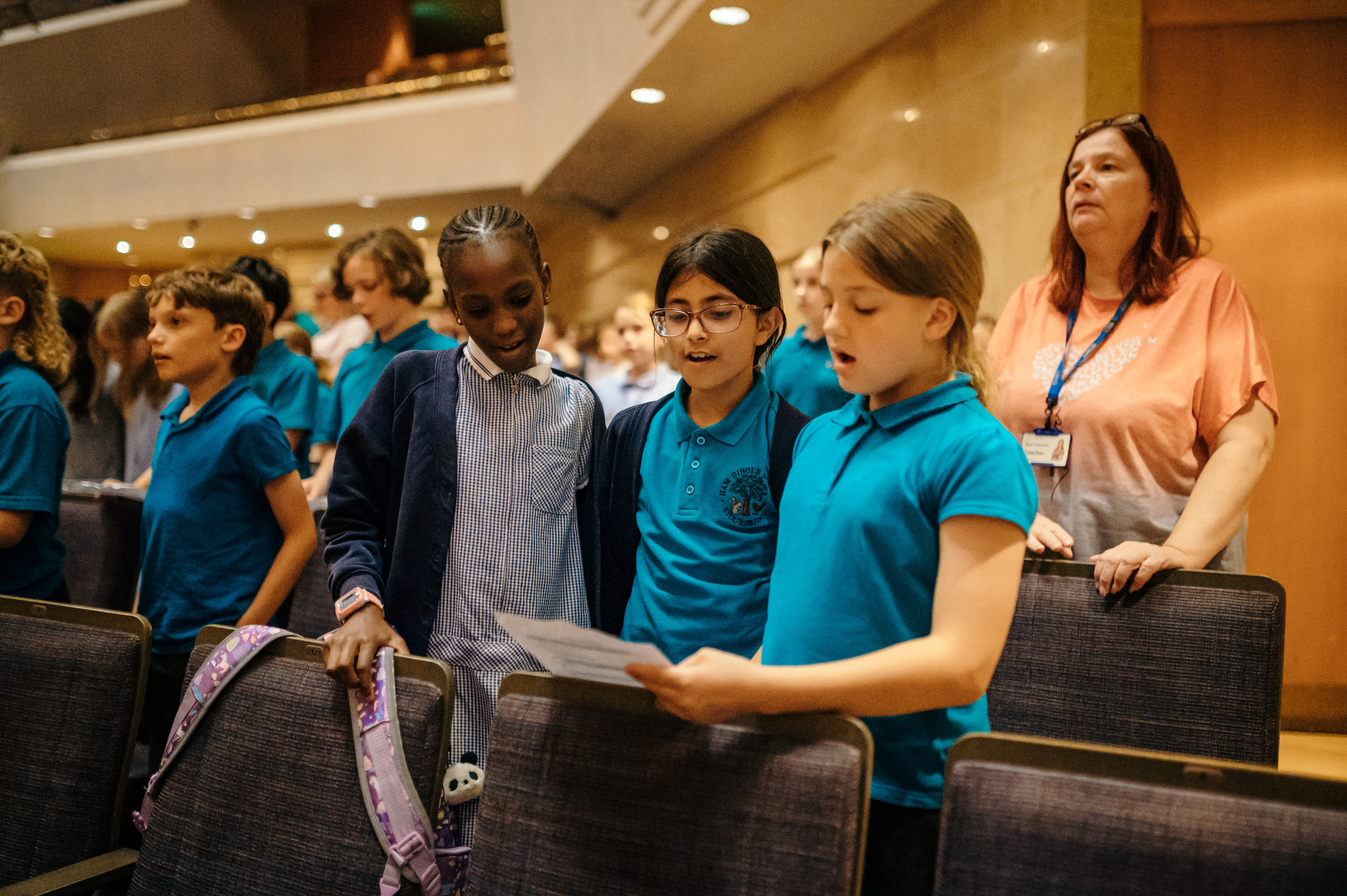 Three girls are singing, reading from one sheet of paper. The girl on the left is wearing a blue check dress and blue cardigan, the middle girl is wearing a turquoise polo shirt, blue cardigan and glasses and the girl on the right is wearing a turquoise polo shirt. They are in a theatre, surrounded by other children.