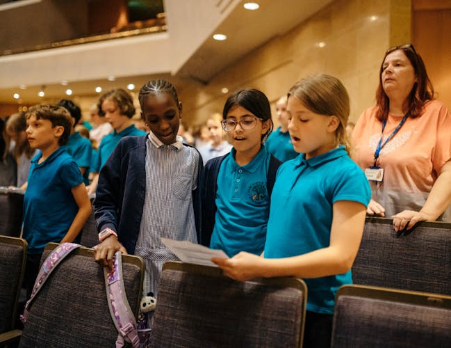 Three girls are singing, reading from one sheet of paper. The girl on the left is wearing a blue check dress and blue cardigan, the middle girl is wearing a turquoise polo shirt, blue cardigan and glasses and the girl on the right is wearing a turquoise polo shirt. They are in a theatre, surrounded by other children.