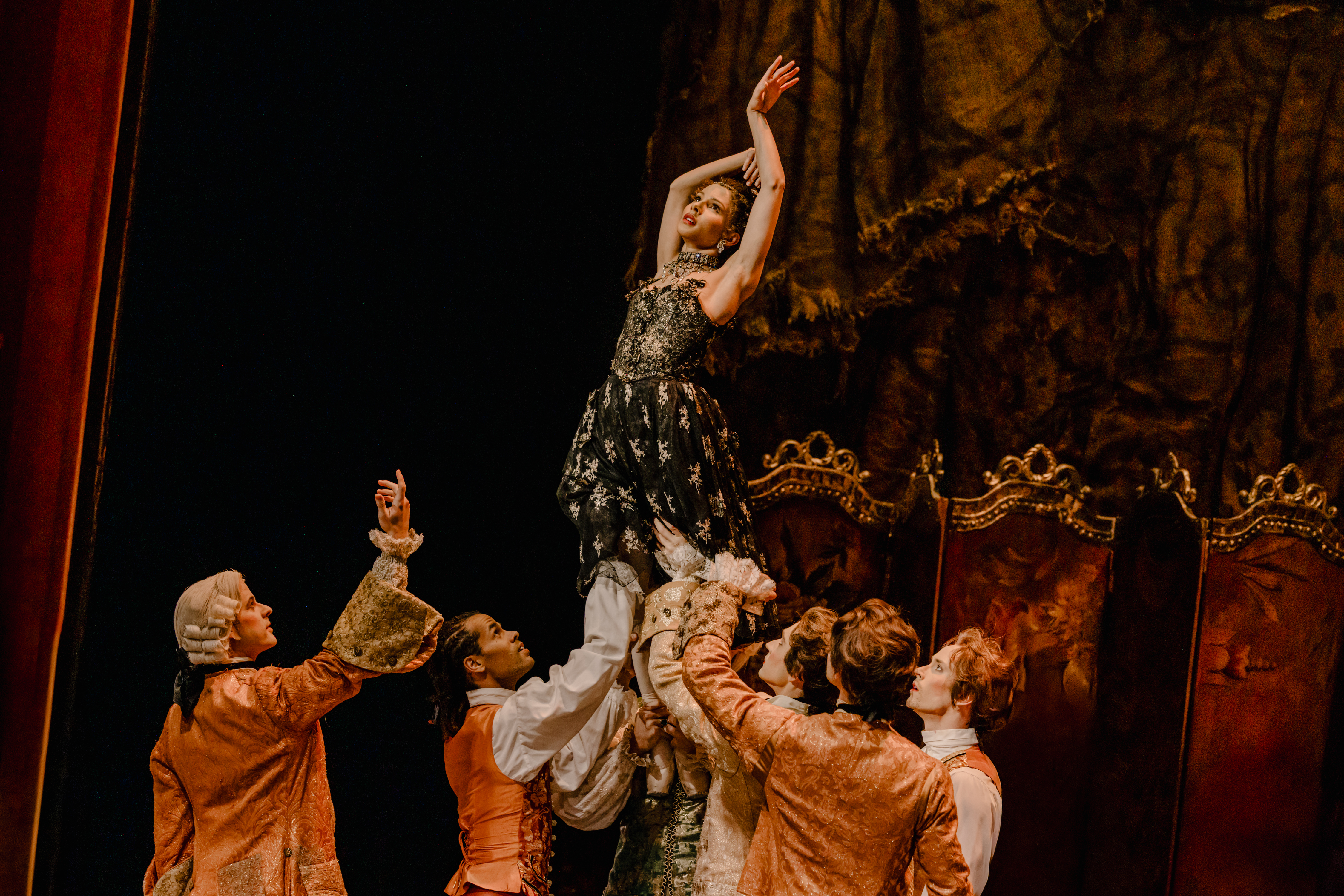 A ballet dancer wearing a black and gold dress is held up by their feet by several other ballet dancers on an elaborate stage. She holds her arms upwards and looks upwards. 