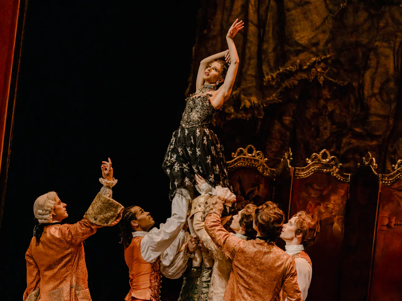 A ballet dancer wearing a black and gold dress is held up by their feet by several other ballet dancers on an elaborate stage. She holds her arms upwards and looks upwards.