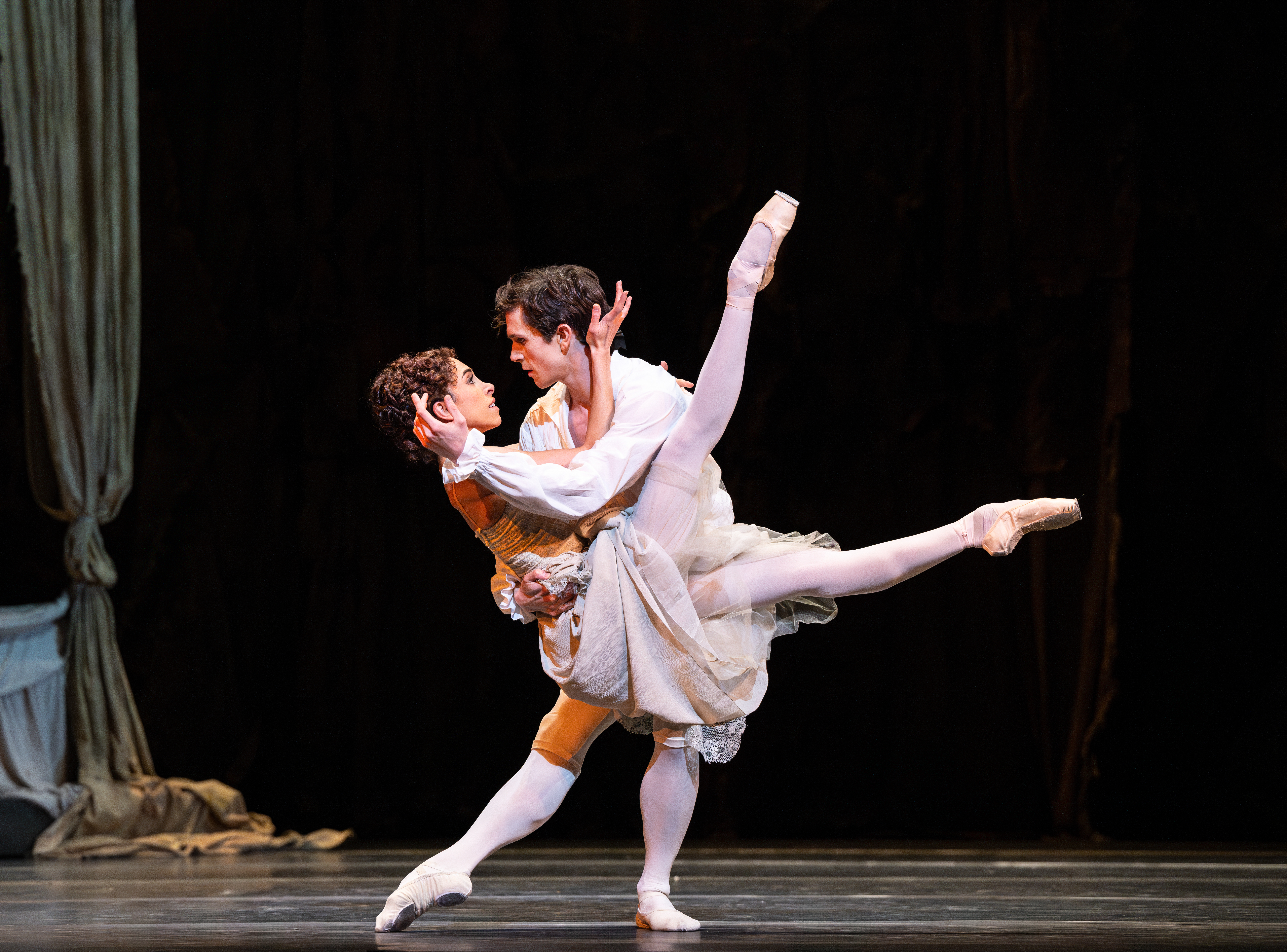 One ballet dancer wearing white lifts another dancer wearing a peasant style dress into the air with her legs raised upwards. (1 of 7)