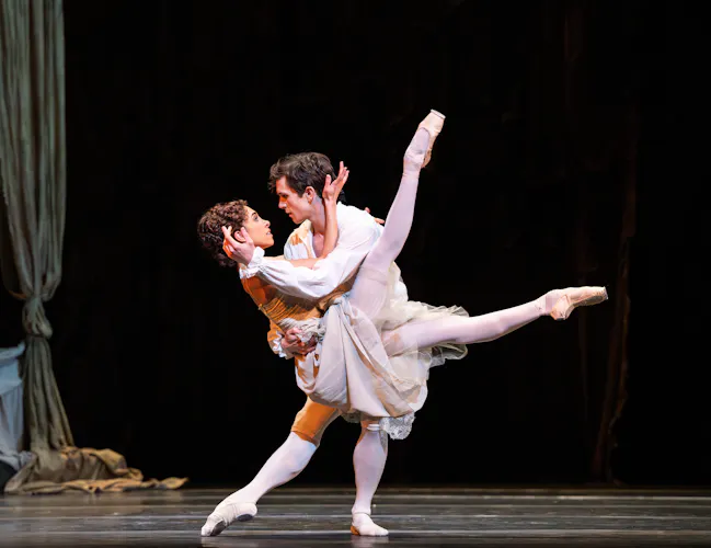 One ballet dancer wearing white lifts another dancer wearing a peasant style dress into the air with her legs raised upwards. (1 of 7)