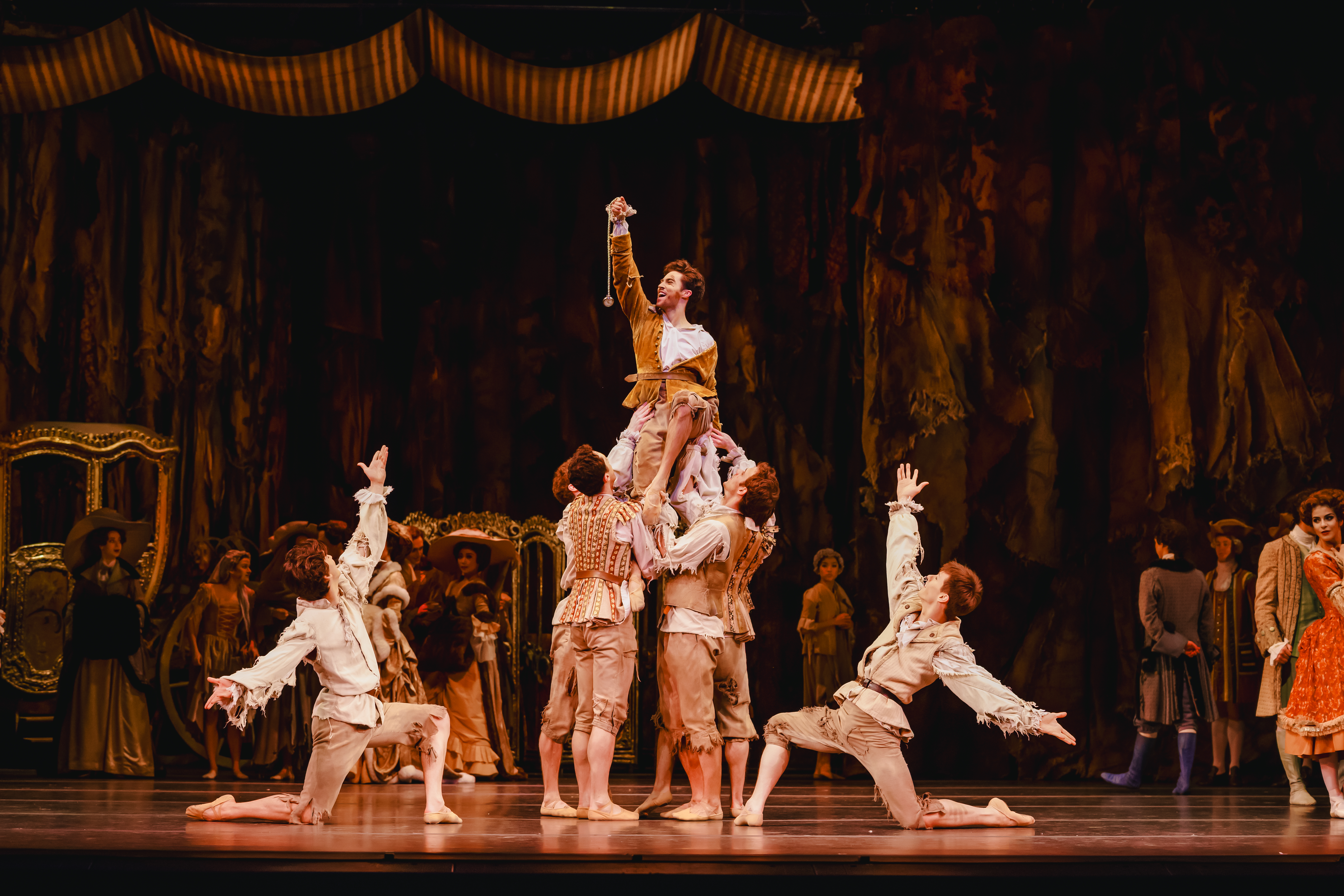 A ballet dancer is held up by several other dancers in the centre of an elaborately decorated stage with other dancers in the background watching on in the performance. They hold their right arm in the air, triumphantly. They are flanked by two dancers who kneel on one knee either side with their arms raised upwards. (3 of 7)