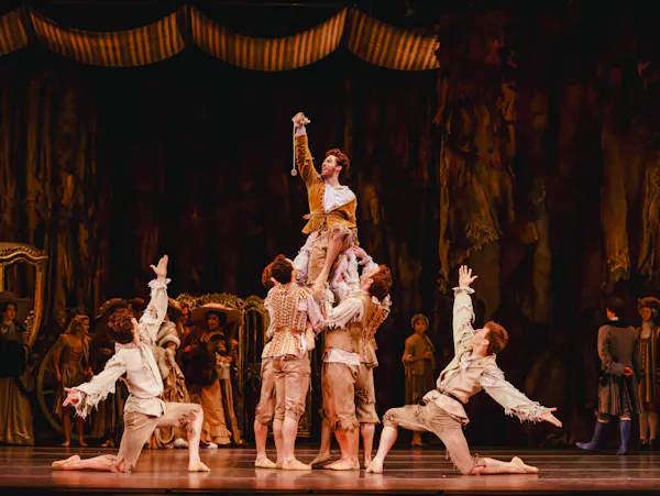 A ballet dancer is held up by several other dancers in the centre of an elaborately decorated stage with other dancers in the background watching on in the performance. They hold their right arm in the air, triumphantly. They are flanked by two dancers who kneel on one knee either side with their arms raised upwards.
