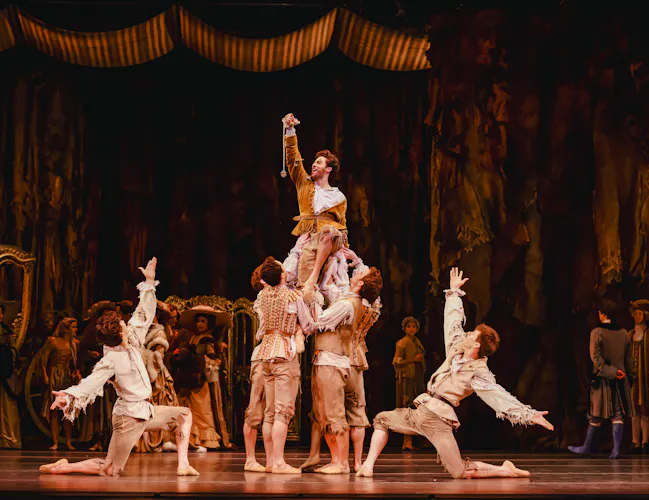 A ballet dancer is held up by several other dancers in the centre of an elaborately decorated stage with other dancers in the background watching on in the performance. They hold their right arm in the air, triumphantly. They are flanked by two dancers who kneel on one knee either side with their arms raised upwards. (3 of 7)