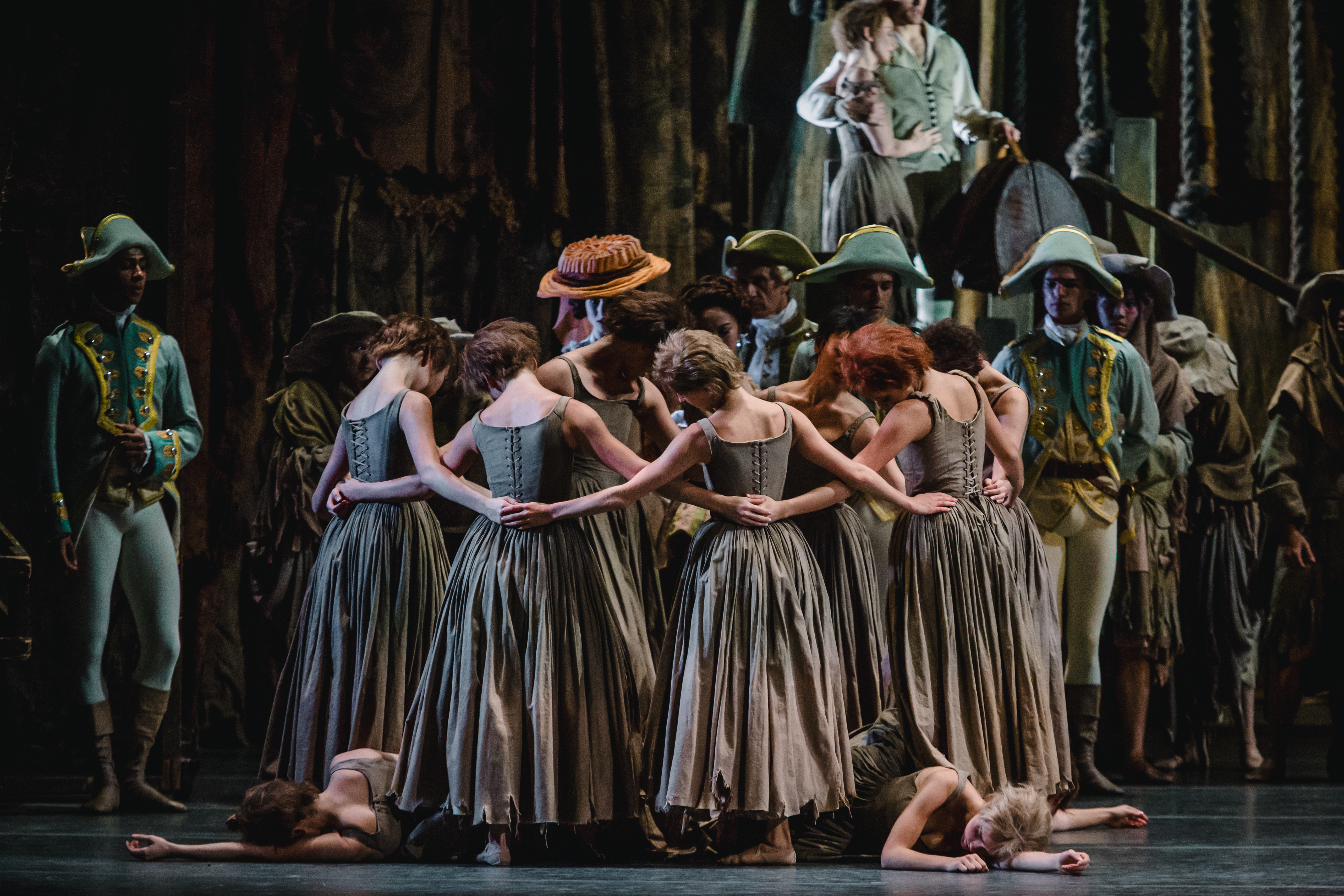 The back of several dancers wearing long grey dresses linking arms in a circle. Two dancers lie at their feet with their arms stretched outwards as performers wearing soldiers uniforms look on. (6 of 7)