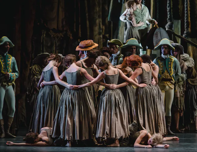 The back of several dancers wearing long grey dresses linking arms in a circle. Two dancers lie at their feet with their arms stretched outwards as performers wearing soldiers uniforms look on. (6 of 7)