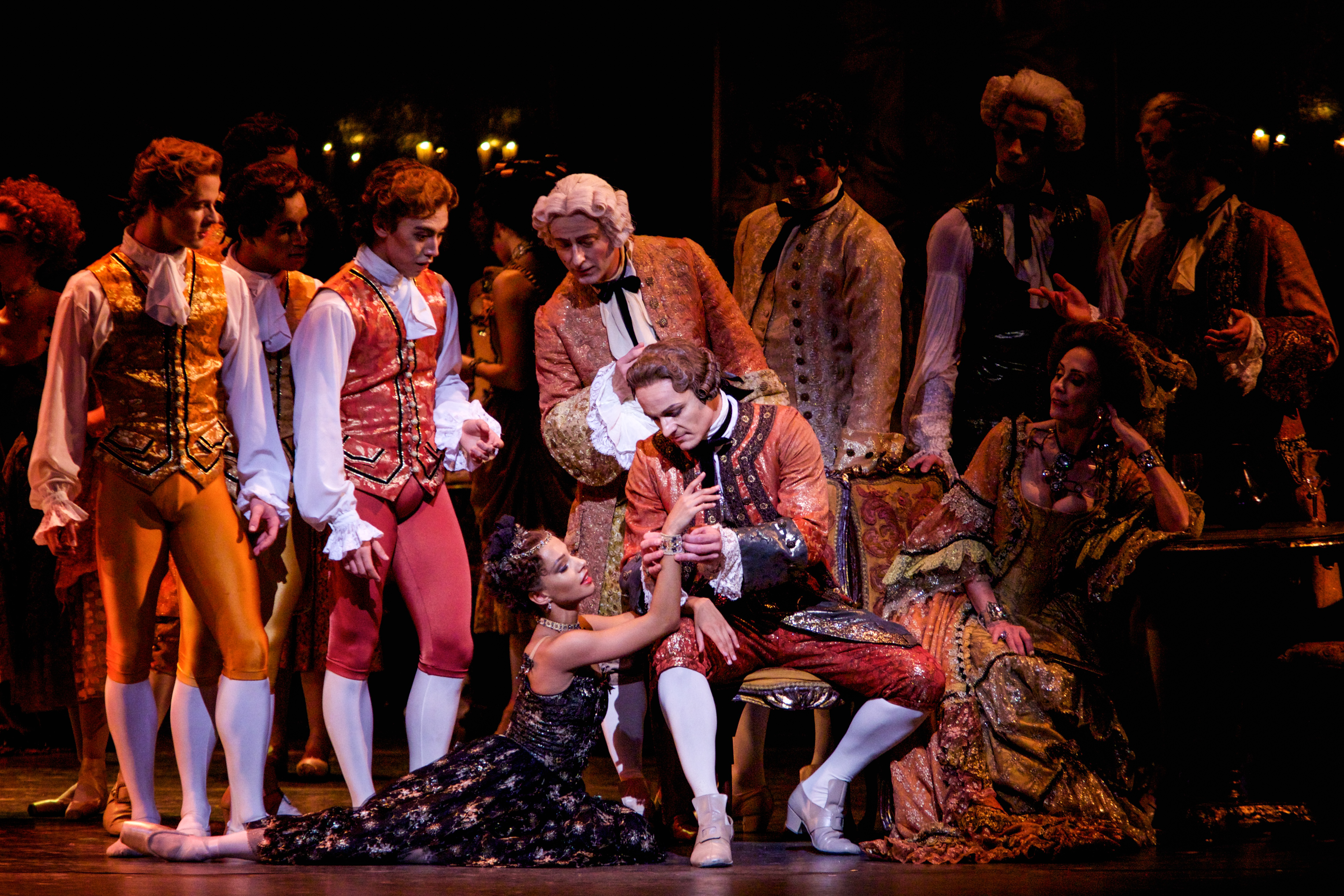A ballet dancer wearing red breeches, white long socks, elaborately embroidered jacket and a powder wig sits on a chair. A dancer wearing a black and gold embroidered dress sits at his feet. Behind them are a group of performers similarly dressed. (2 of 7)
