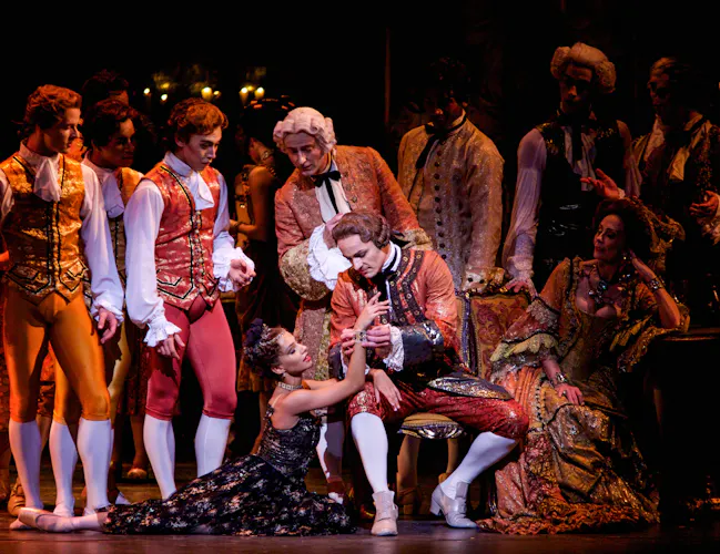A ballet dancer wearing red breeches, white long socks, elaborately embroidered jacket and a powder wig sits on a chair. A dancer wearing a black and gold embroidered dress sits at his feet. Behind them are a group of performers similarly dressed. (2 of 7)