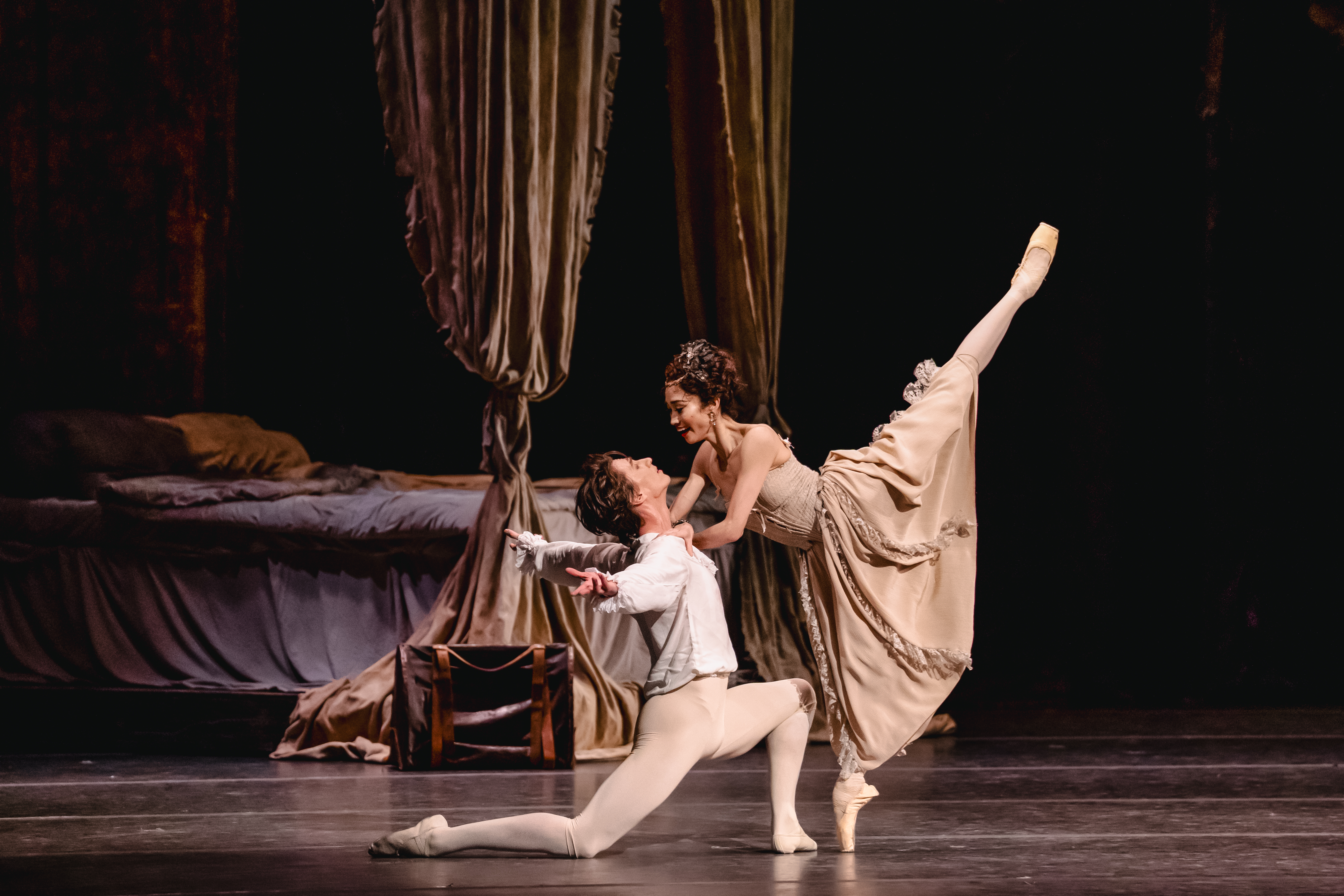Two ballet dancers embrace passionately on a stage. Behind them is a four poster bed draped with luxurious curtains and unmade sheets. (5 of 7)