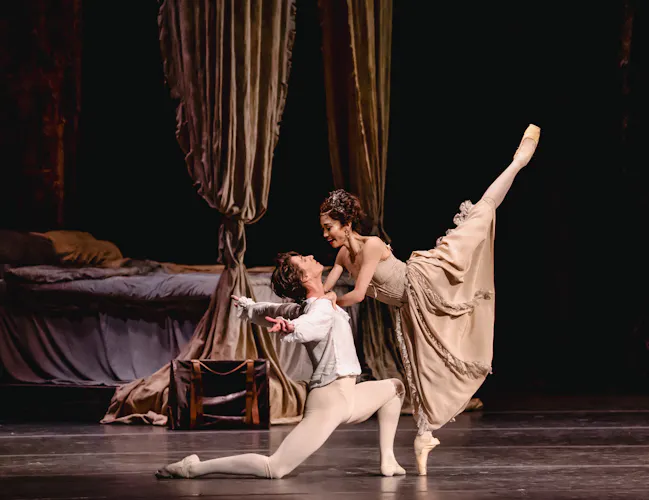 Two ballet dancers embrace passionately on a stage. Behind them is a four poster bed draped with luxurious curtains and unmade sheets. (5 of 7)