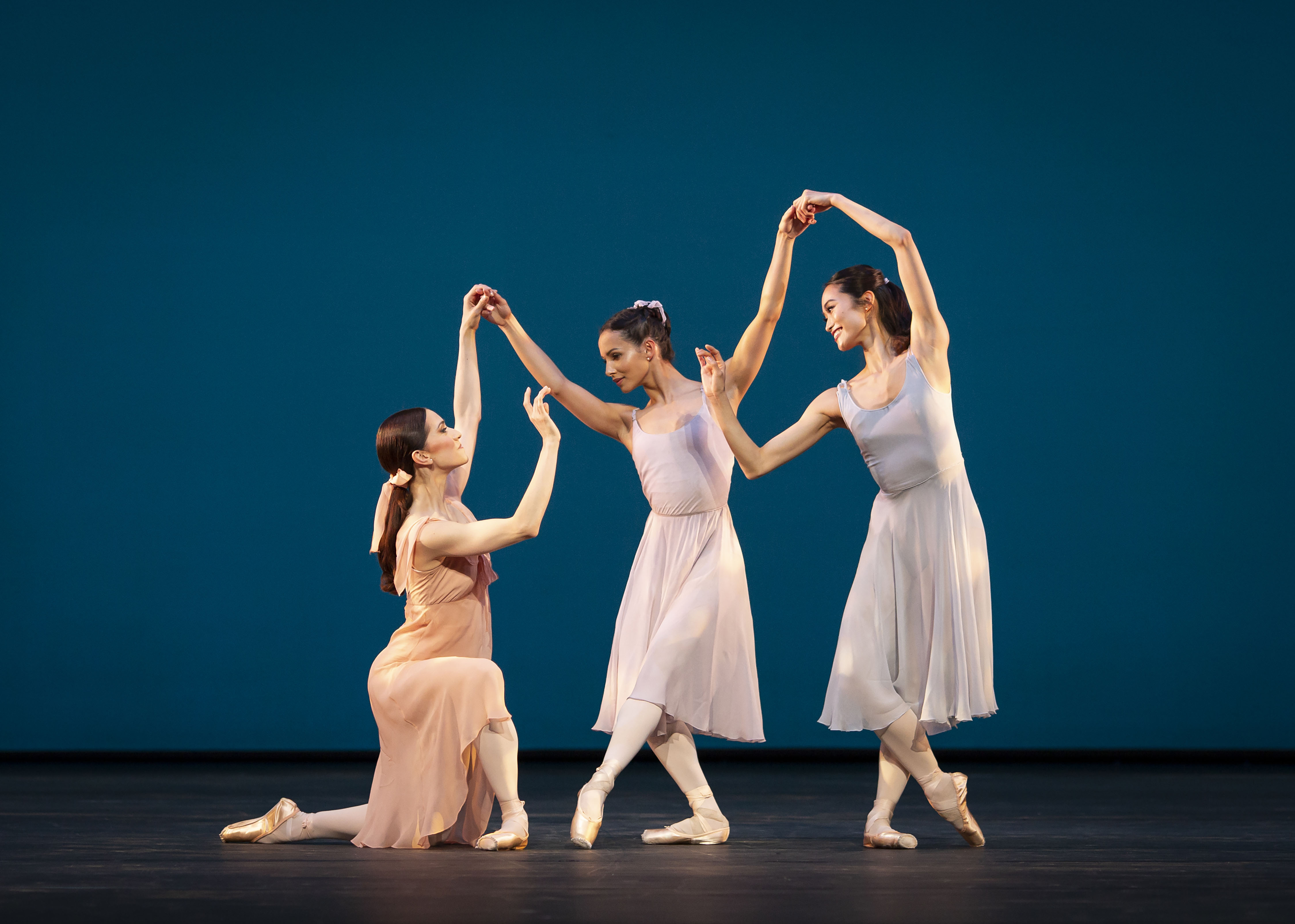 Three ballet dancers perform together wearing light coloured dresses in front of a blue backdrop on a stage. 