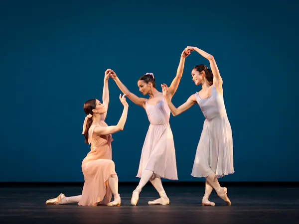 Three ballet dancers perform together wearing light coloured dresses in front of a blue backdrop on a stage.