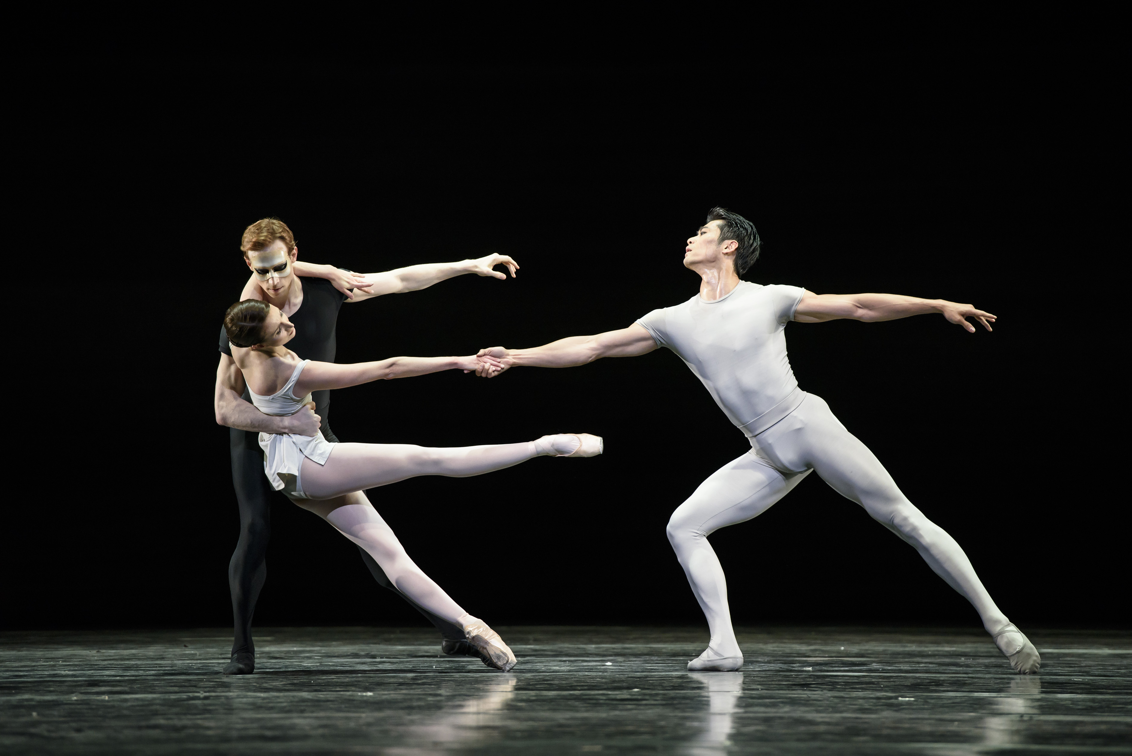 Three ballet dancers perform together on stage: two are embracing while the other dancer stretches outwards and holds onto one of the dancers outstretched hand. (2 of 6)