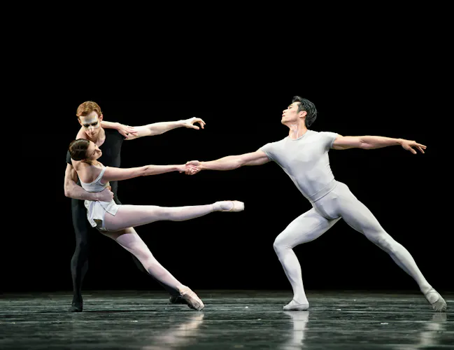 Three ballet dancers perform together on stage: two are embracing while the other dancer stretches outwards and holds onto one of the dancers outstretched hand. (2 of 6)