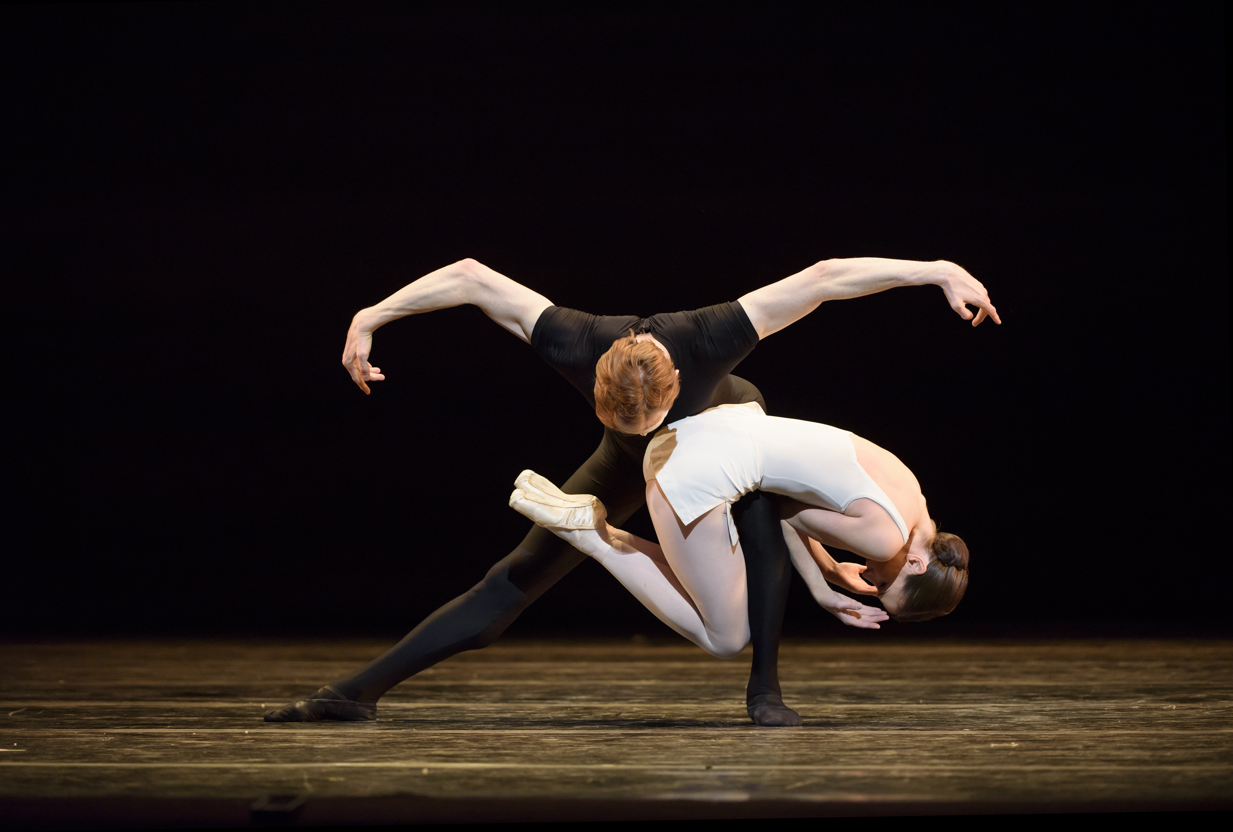 Two ballet dancers perform together on stage. One wears all black and stands with their legs wide apart. The other dancer wearing a short white dress lies on the other dancers bent leg creating an interesting shape. (4 of 6)