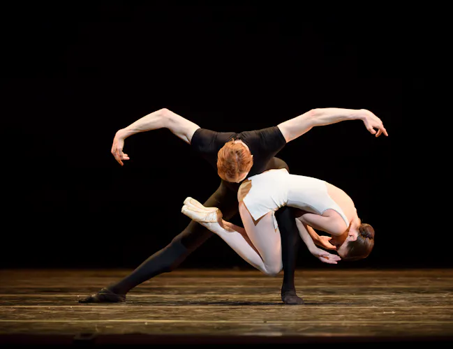 Two ballet dancers perform together on stage. One wears all black and stands with their legs wide apart. The other dancer wearing a short white dress lies on the other dancers bent leg creating an interesting shape. (4 of 6)