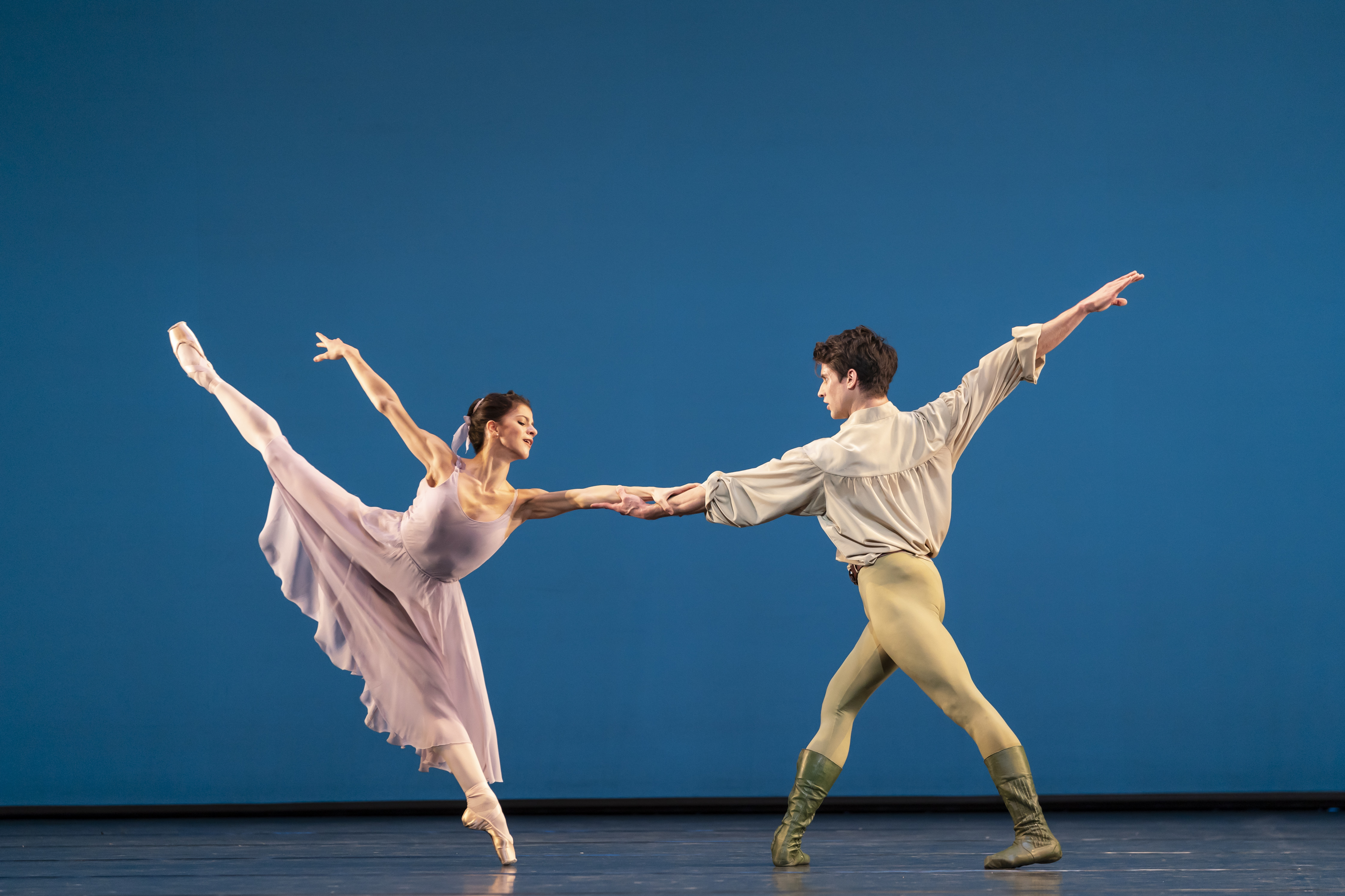 Two ballet dancers perform together on a stage with a bright blue backdrop. One wears a long pink dress and the other is all in white. (1 of 6)