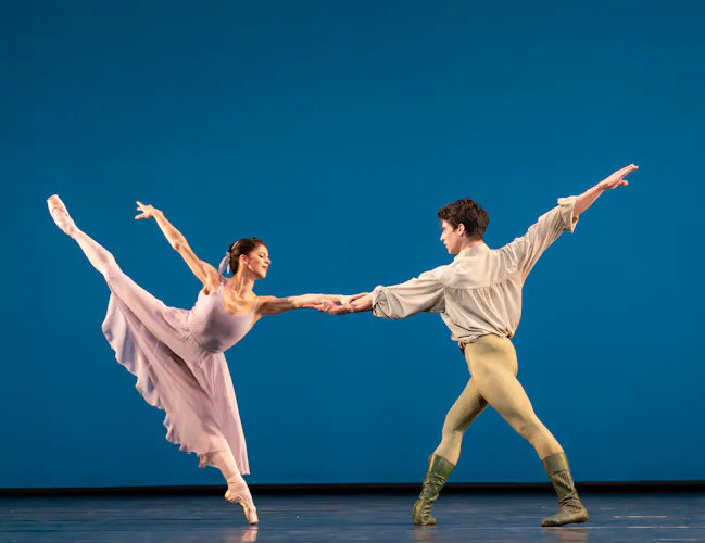 Two ballet dancers perform together on a stage with a bright blue backdrop. One wears a long pink dress and the other is all in white. (1 of 6)