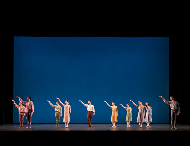 Several ballet dancers stand on a stage with a blue backdrop. They all hold their right arm upwards. (5 of 6)