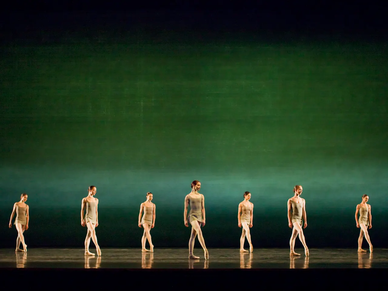 Several ballet dancers stand en pointe on a stage lit with a gradient green backdrop.