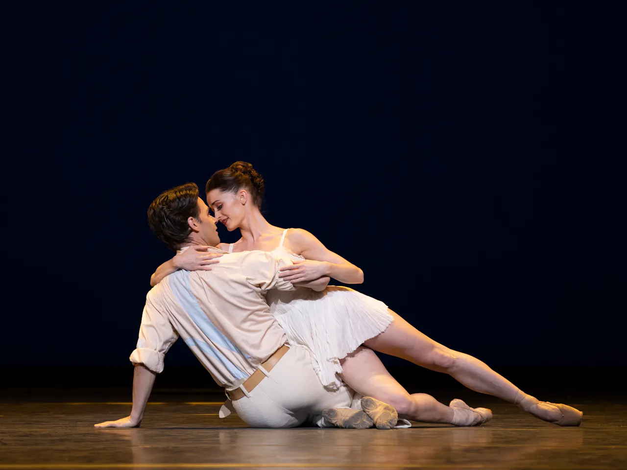 Two ballet dancers lie in a passionate embrace on the floor of a stage with a dark background. 
