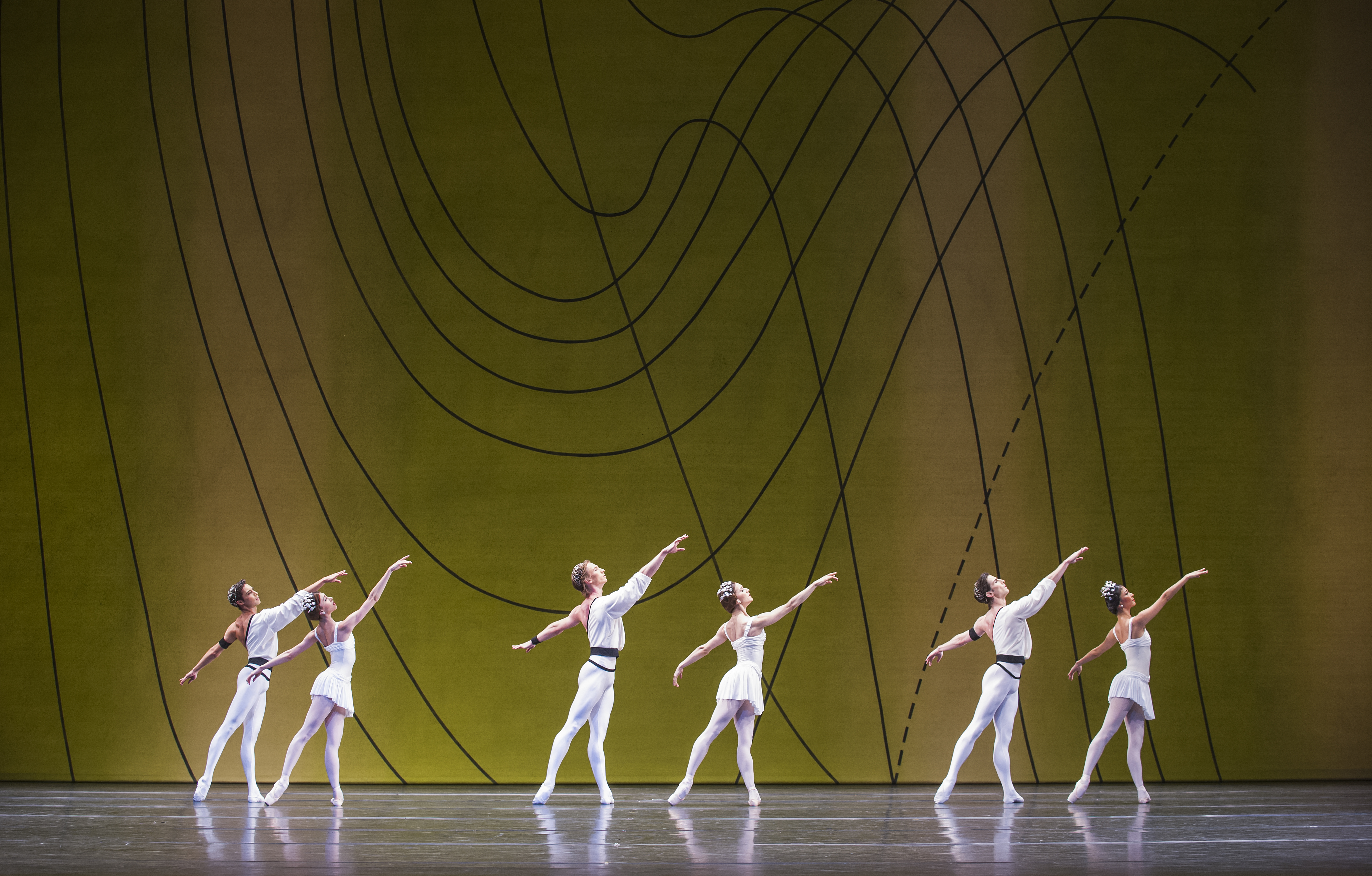 Several ballet dancers perform on stage, all are wearing white costumes. They perform in front of a yellow and green gradient backdrop with flowing geometric patterns.