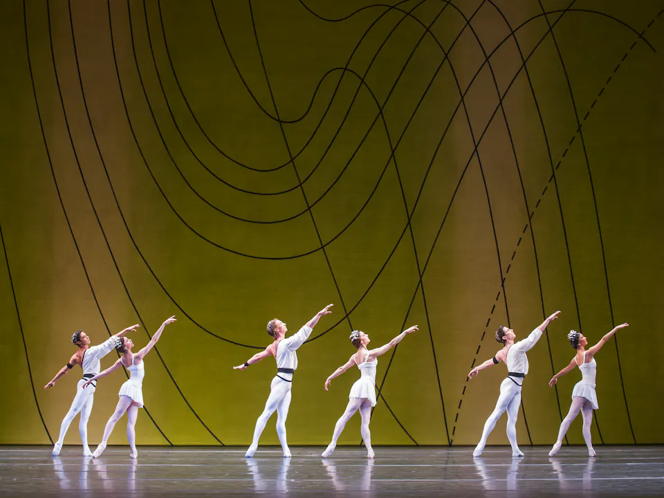 Several ballet dancers perform on stage, all are wearing white costumes. They perform in front of a yellow and green gradient backdrop with flowing geometric patterns.