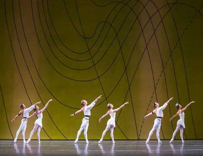 Several ballet dancers perform on stage, all are wearing white costumes. They perform in front of a yellow and green gradient backdrop with flowing geometric patterns. (3 of 10)