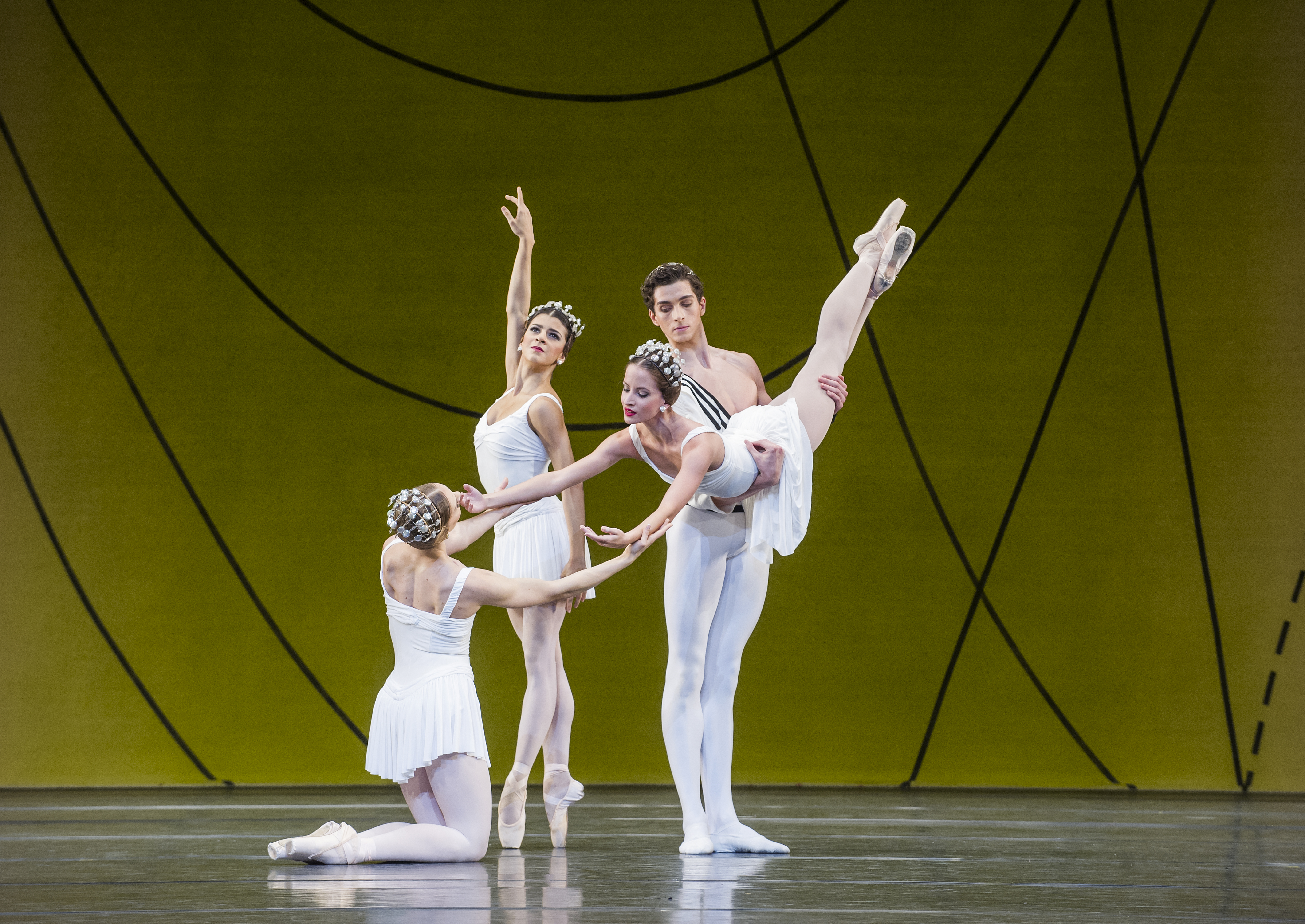 Several ballet dancers perform on stage, all are wearing white costumes. They perform in front of a yellow and green gradient backdrop with flowing geometric patterns. (5 of 6)