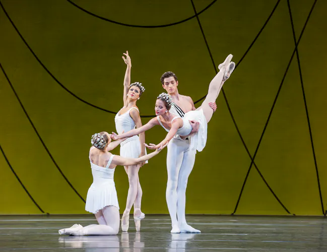 Several ballet dancers perform on stage, all are wearing white costumes. They perform in front of a yellow and green gradient backdrop with flowing geometric patterns. (5 of 6)