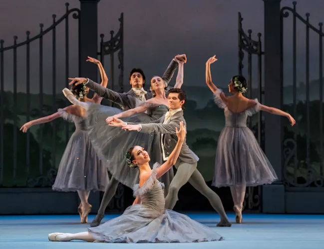Several ballet dancers wearing grey dresses with flowers in their hair perform with dancers wearing grey suits. They perform in front of an ornate gate with a painterly rural background. (3 of 6)